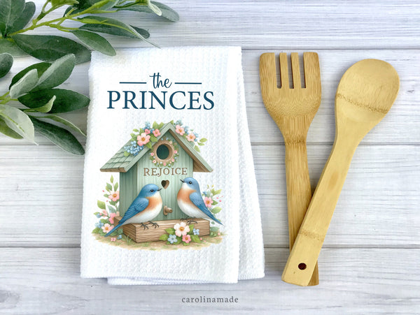 Teal bird house with two birds and flowers surrounding it. The top is personalized with family name. On white waffle weave towel.