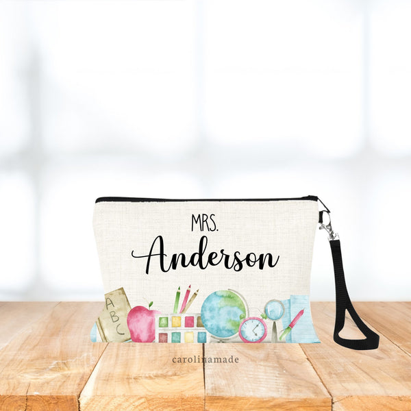 Watercolor Teacher border design along the bottom of the bag. Paints, paper, pend, clock, apple, pencils, globe. Above it is personalized with teachers name.  Linen wristlet bag includes black strap.