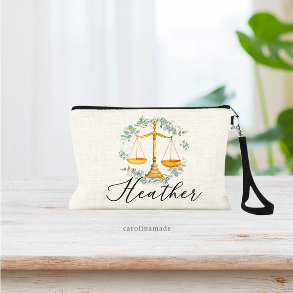 This natural linen wristlet features the scales of justice in the center of the bag in watercolor gold design. With a watercolor greenery wreath surrounding it. A name is personalized in a beautiful script under the wreath.
