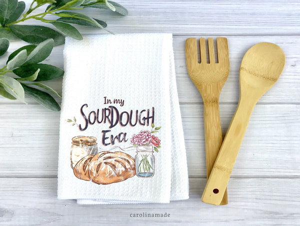 This white waffle weave towel says In my sourdough era and shows a jar of dough, bread, and a jar of flowers. The perfect kitchen towel for bakers.