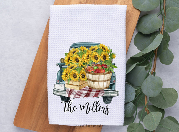 This white waffle weave towel features a truck bed full of sunflowers and a basket of apples. It is personalized at the bottom with your family name.