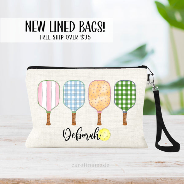 This linen zipper pouch with lining and wristlet strap features four colorful pickleball paddles with various designs on them all in a row. At the bottom it is personalized with your name next to a pickelball ball.