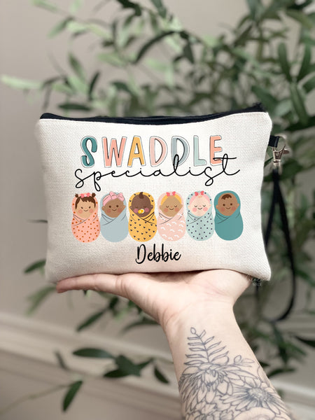 This linen zipper pouch features the cutest little babies swaddled in a row with the text Swaddle specialist above them. A name is personalized at the bottom.