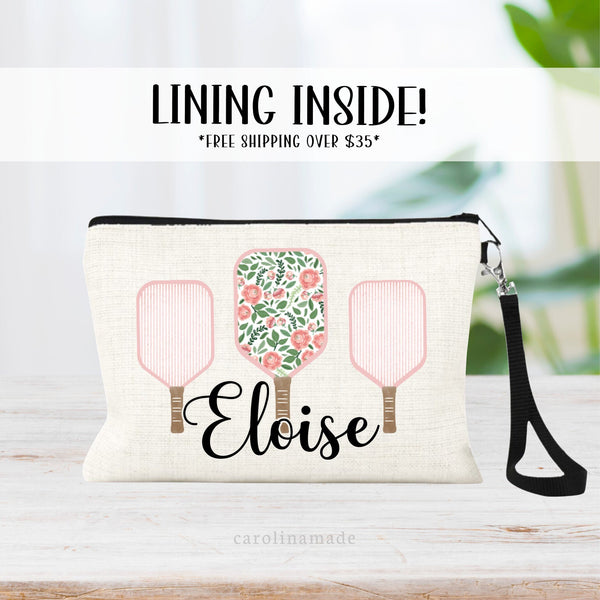 This linen cosmetic bag features 3 pickleball paddles, two with pink stripes and one with pink flowers and greenery. The bottom is personalized with your name.