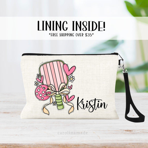 This linen wristlet features a fun girly design with a pickleball paddle, ball, bow and ribbon, hearts and a daisy. It is personalized next to the paddle with your name.