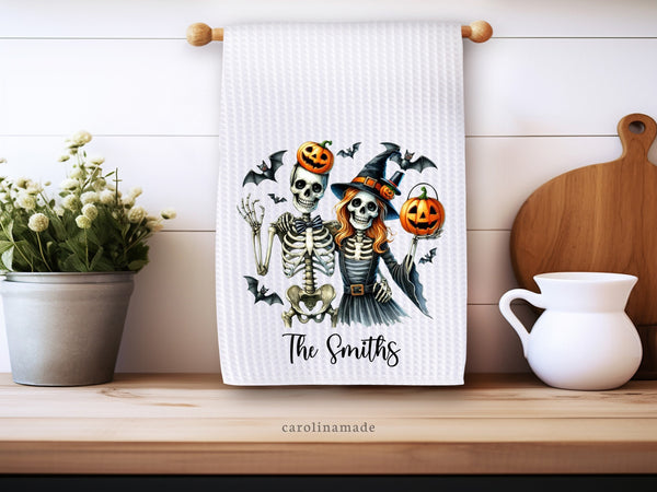 This white waffle weave towel features a skeleton couple dressed in halloween costumes with bats and pumpkins surrounding. It is personalized at the bottom with your family name.