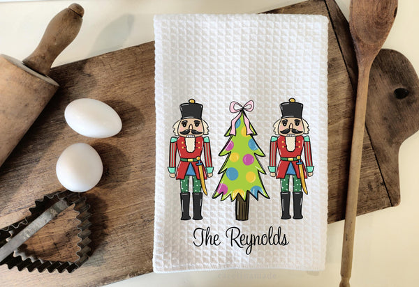 This white waffle weave towel features two nutcrackers with a christmas tree in the center. It is personalized with your family name at the bottom.