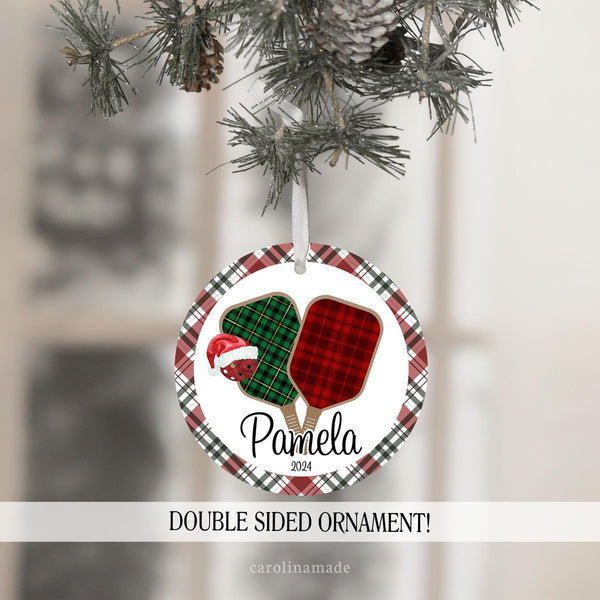 This Christmas ornament features a red & green plaid background with plaid pickleball paddles in the center and a pickleball with santa hat. It is personalized at the bottom with your name and year.