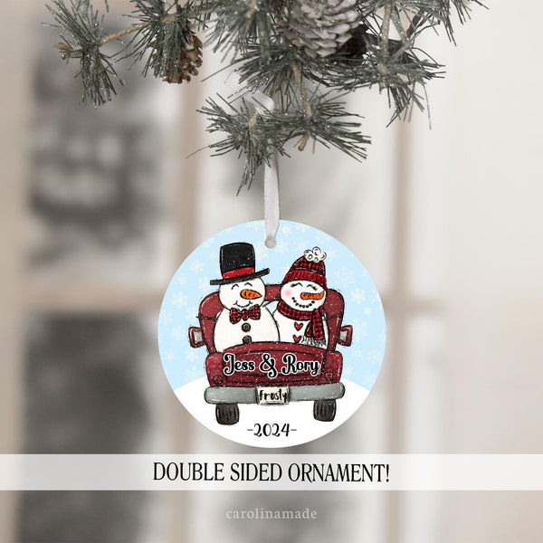 This ornament features a Mr and Mrs Snowman In the back of a truck with a snowy background. It is personalized on the truck with their name and at the bottom with the year.