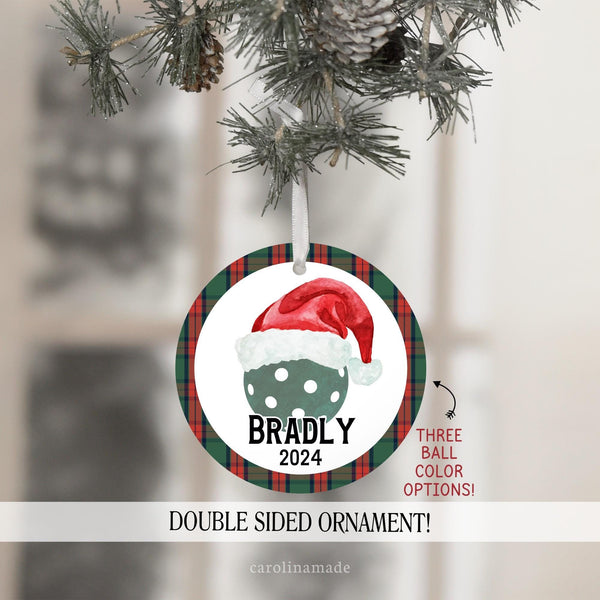 This Christmas ornament features a red and green plaid background with a large pickleball in the center with a santa hat on it. It is personalized at the bottom with your name and year. Choose green or red ball.