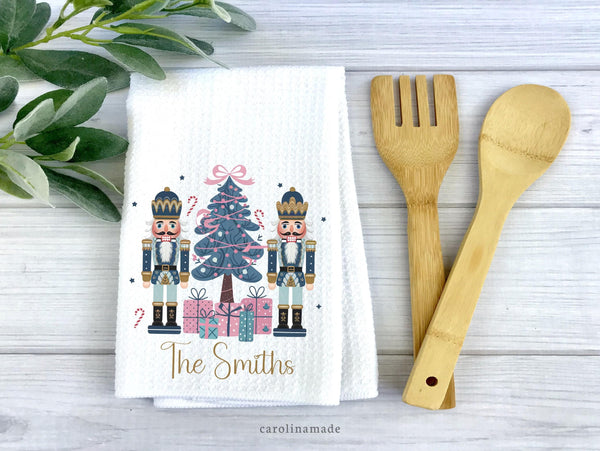 This white waffle weave hand towel features two nutcrackers and a blue christmas tree with pink gifts. It is personalized at the bottom with your family name.