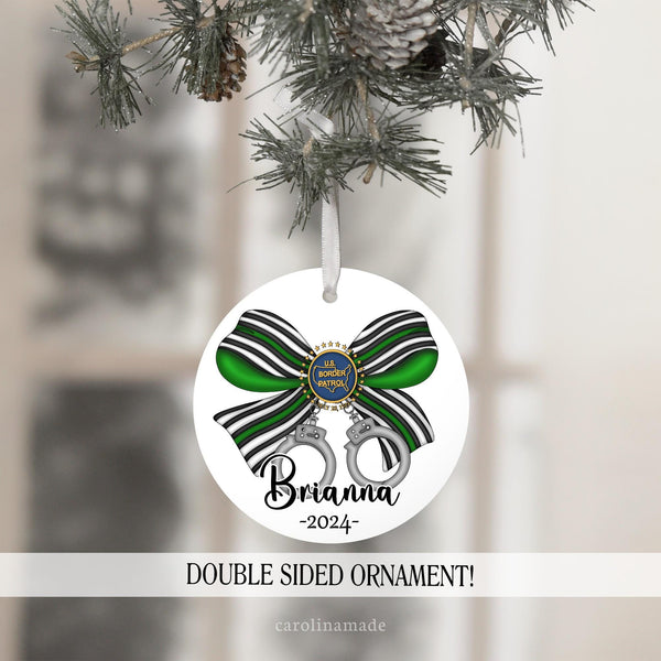 This ornament features a large bow in border patrol colors with hand cuffs handing off of it. It is personalized at the bottom with name and the year.