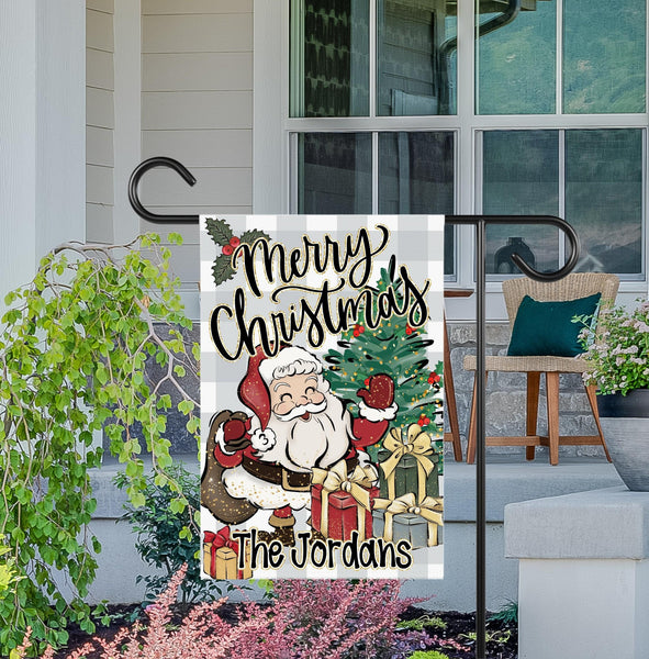 This garden flag features a gray plaid background. Santa is waving on the front with a Christmas tree and gifts. The top says Merry Christmas and the bottom is personalized with your family name.