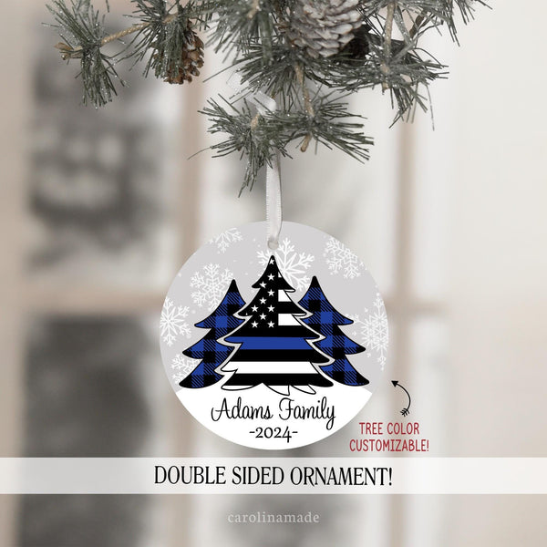 This ornament features gray snowy background with three Christmas trees in the center. Two with blue plaid. And the center large tree is a flag with a thin blue line. It is personalized at the bottom with your name and the year.