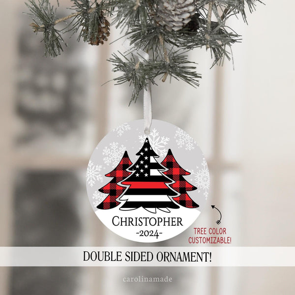 This ornament features gray snowy background with three Christmas trees in the center. Two with red plaid. And the center large tree is a flag with a thin red line. It is personalized at the bottom with your name and the year.