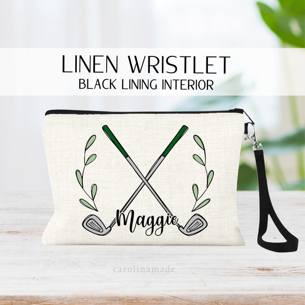 This linen zipper pouch features a soft black lining on the inside with black zipper and black wristlet strap. The front has a crossed golf clubs with wreaths and green leaves around it. It is personalized with name at the bottom.