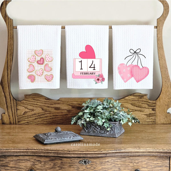 This set of 3 waffle weave towels features a beautiful pink valentine's day decor set. Cookies with icing and sprinkles, pink hearts and a calendar marking February 14th and pink flowers.