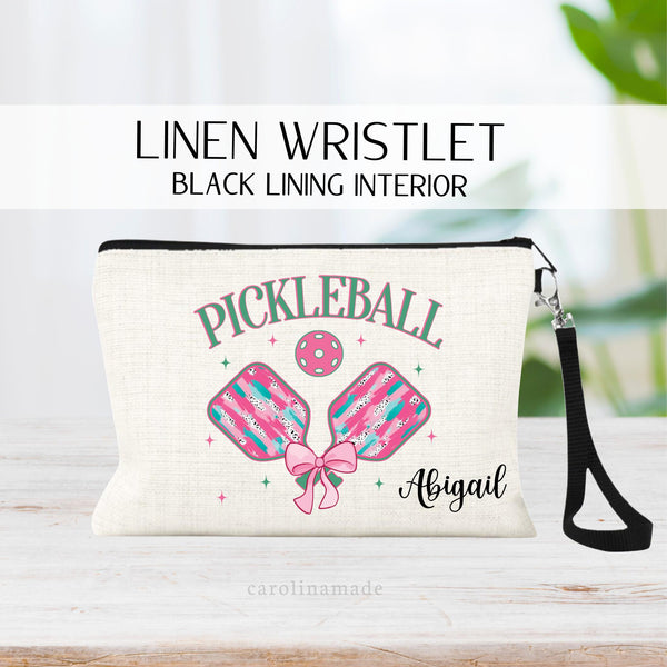 This linen zipper pouch is personalized for a pickleball player. It says Pickleball at the top in pink and green letters. Then two pickleball paddles with brush stroke design and pink ball with a coquette bow on the paddles.