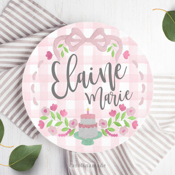 This 10" Plate features a pink gingham background with a grand millennial design with bow at the top, flowers and a cake at the bottom. The center is personalized with your childs name.