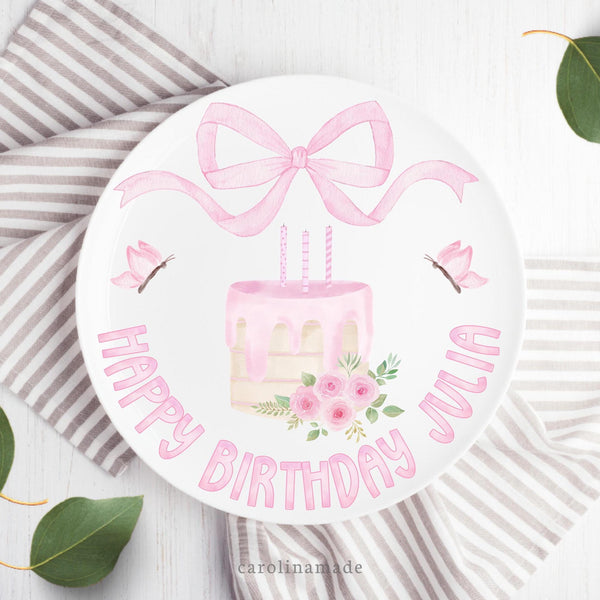 This white 10 inch plate features a pink watercolor bow at the top, a cake with candles, flowers and leaves in the center, butterflies on both sides. and says Happy Birthday at the bottom with your personalized name.