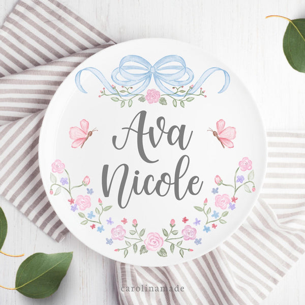 This white 10 inch plate features a blue watercolor bow at the top with greenery and flowers, your personalized name in the center, butterflies and greenery and flowers at the bottom.