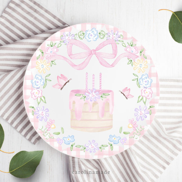 This white 10 inch plate features a pink watercolor bow at the top, a cake with candles, flowers and leaves in the center, butterflies on both sides. There is a floral wreath and pink gingham around the sides.