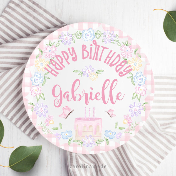 This white 10 inch plate features a pink watercolor bow at the top, a cake with candles, flowers and leaves in the center, butterflies on both sides. There is a floral wreath and pink gingham around the sides. Personalized with name in center.