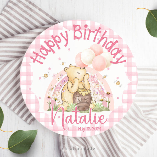 This ten inch plate features a pink gingham scallop edge with pink pokadots in the background. The top says happy birthday with a classic pooh art in the center. There is a rainbow, flowers, bees and balloons around Pooh. The bottom is personalized with name and date of birth.