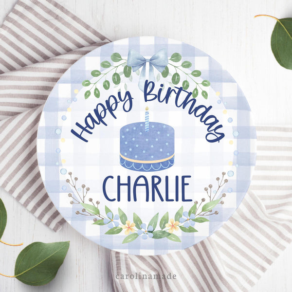 This white 10 inch plate features a blue gingham background with a blue bow and a floral blue and yellow wreath with greenery. The center says Happy Birthday with a blue cake in the center and your childs name at the bottom.