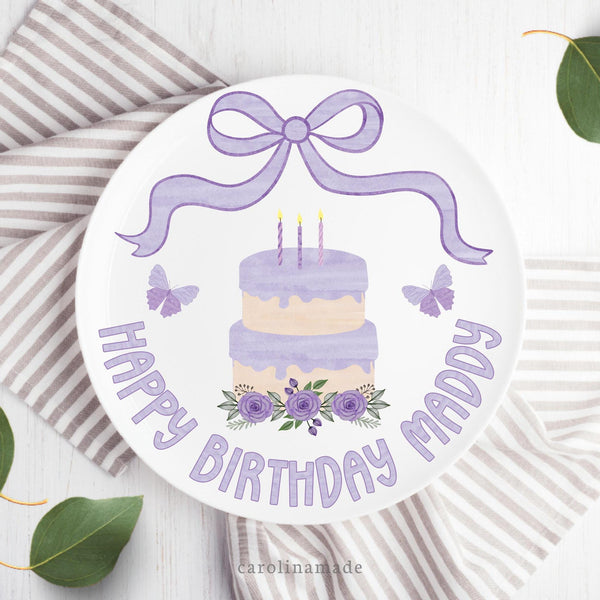 This white 10 inch plate features a purple watercolor bow at the top, a cake with candles, flowers and leaves in the center, butterflies on both sides. and says Happy Birthday at the bottom with your personalized name.