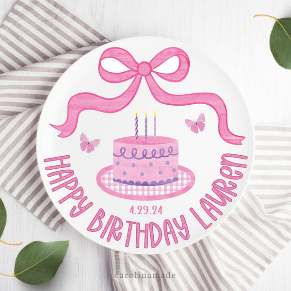 This white 10 inch plate features a pink watercolor bow at the top, a cake with candles, and butterflies on both sides. and says Happy Birthday at the bottom with your personalized name.