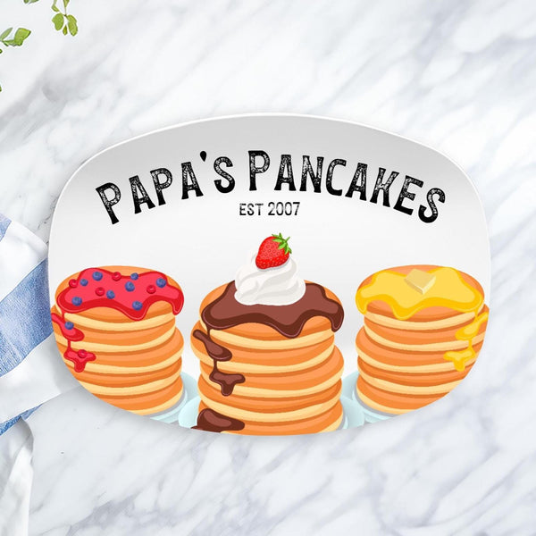 This white serving tray features three stacks of pancakes at the bottom. One with strawberry syrup, one with chocolate syrup whip creak and strawberry and the other with butter syrup.  The top says Papas Pancakes and can be personalized to whomever youd like.  And the established date under text.