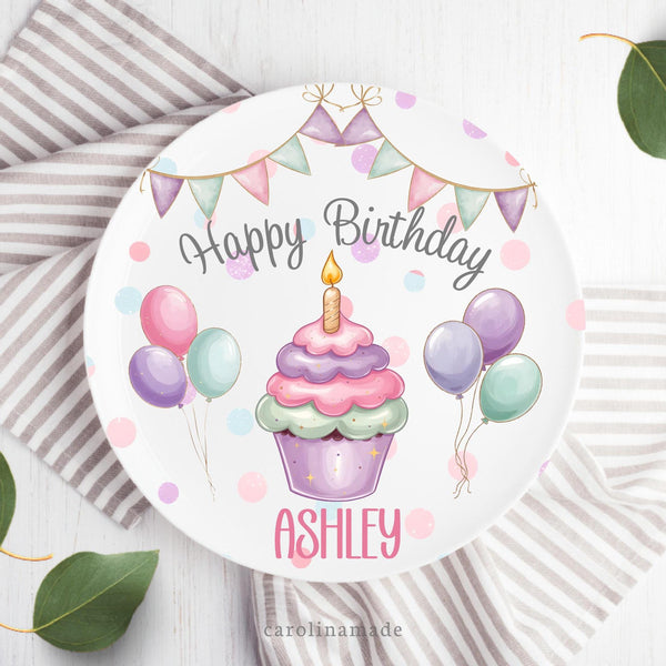 This white 10 inch plate features a pink purple blue poka dot background with pastel banner at the top, a large cupcake in the center with candle and balloons around it. The plate says Happy Birthday in cursive and says your personalized name at the bottom.