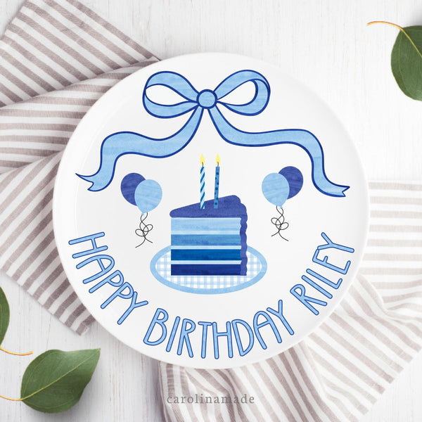 This 10 inch plate features white background and blue theme birthday design. A blue coquette bow at the top with a multi layer cake in the center in different shades of blue. The bottom says Happy birthday and is personalized with your childs name. The plate also features balloons in shades of blue.