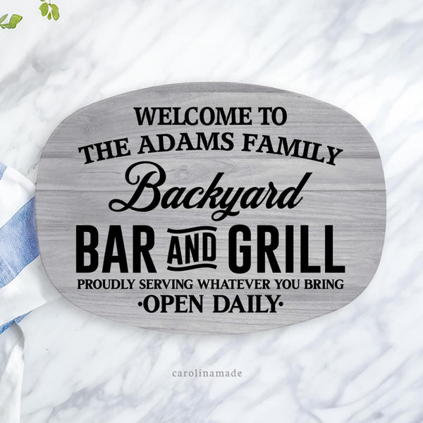 This 10 x 14 serving platter features a light gray wood grain background with black text over it. It says Welcome to the ___ family backyard bar and grill. Proudly serving whatever you bring. Open daily. And is personalized with a family name.