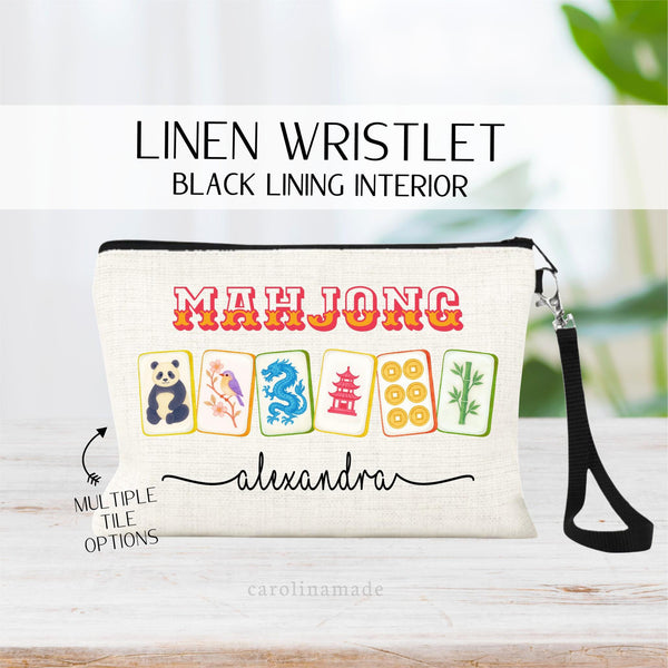 May include: A white linen wristlet with a black lining interior. The wristlet has a black strap and features a colorful Mahjong tile design with the text 'Mahjong' and 'alexandra' printed on it.