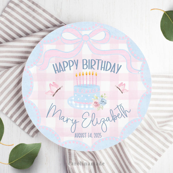 This white 10 inch plate features a pink and blue watercolor bow at the top, blue scallop edges, and pink gingham background, a cake with candles, flowers and leaves in the center, butterflies on both sides. and says Happy Birthday at the bottom with your personalized name and date of birth.