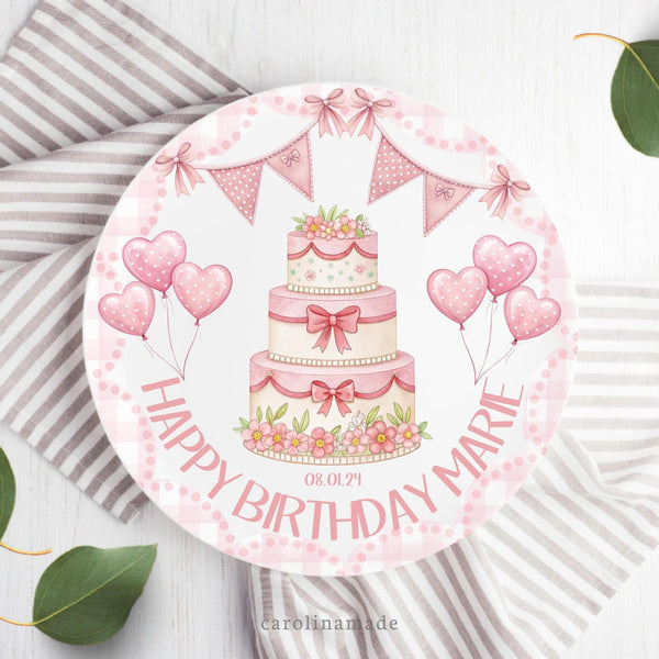 This girls birthday plate features a pink gingham scallop border. There are pink banners, pink balloons, Pink dots, and a cake in the center. The bottom text says happy birthday and is personalized with your daughter's name.