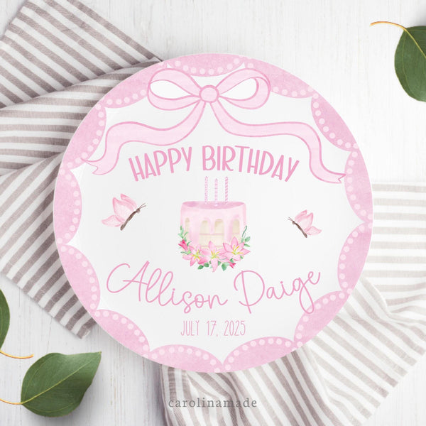 This white 10 inch plate features a pink watercolor bow at the top, pink scallop edges, a cake with candles, flowers and leaves in the center, butterflies on both sides. and says Happy Birthday at the bottom with your personalized name and date of birth.
