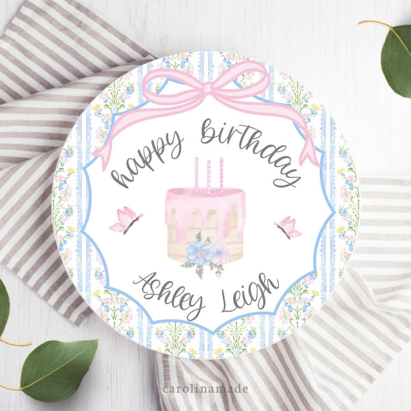 This white 10 inch plate features a pink watercolor bow at the top, a cake with candles, flowers and leaves in the center, butterflies on both sides. There is a floral scallop border around the edge. Personalized with name in center.