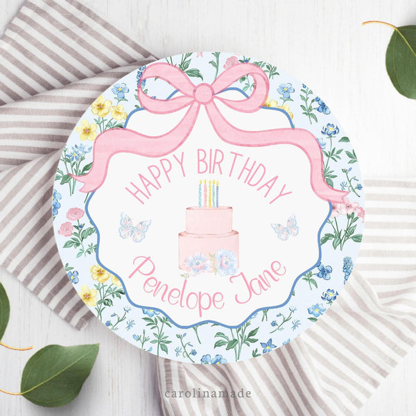 This girls birthday plate features a pink bow at the top with a floral scallop border. The center says Happy birthday and your personalized name. A cake is in the center with flowers and floral butterflies on both sides.